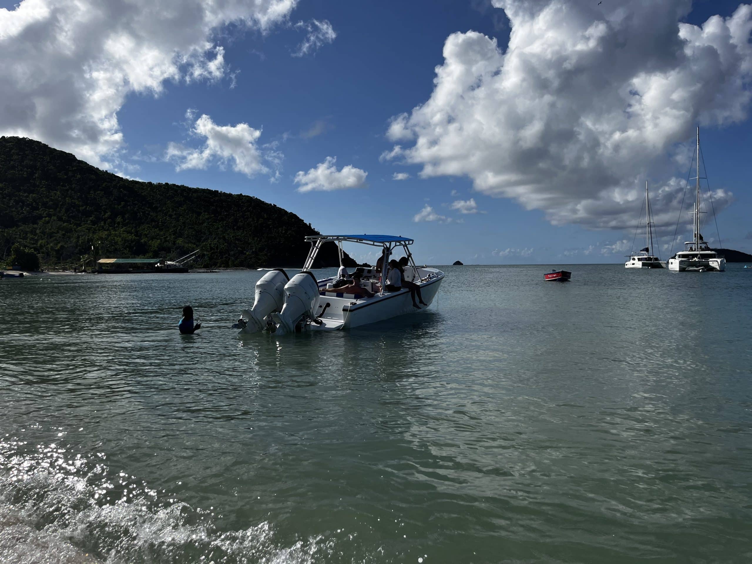 A small motorboat with people on board floats near the shore of a calm bay, surrounded by green hills and anchored sailing boats—an inviting scene for power boat excursions from Bequia under a partly cloudy sky.