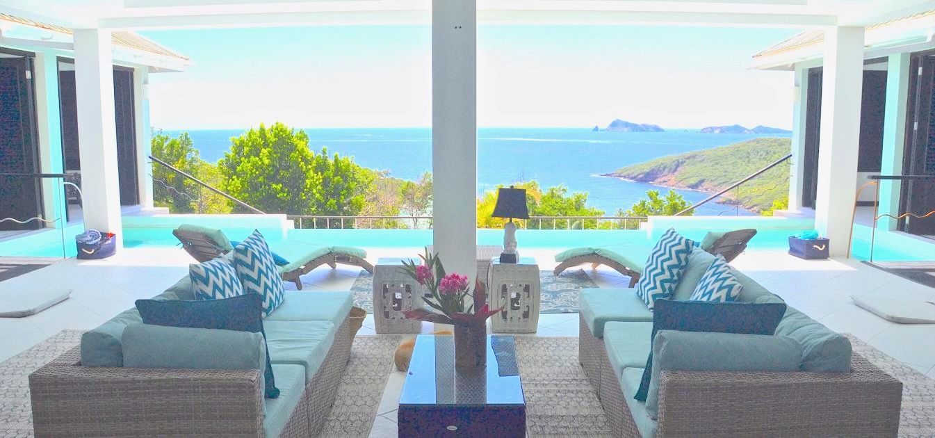 Luxury villa Bequia offers an open-air living space with wicker sofas and blue-patterned cushions, overlooking an infinity pool and a scenic sea view with distant green islands under a bright, clear sky. Photo