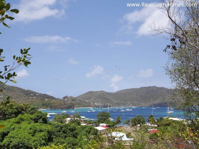 A view of a coastal village with lush green trees, blue sea, anchored sailing boats, and hills in the background under a partly cloudy sky. Website text appears in the top right corner. Photo