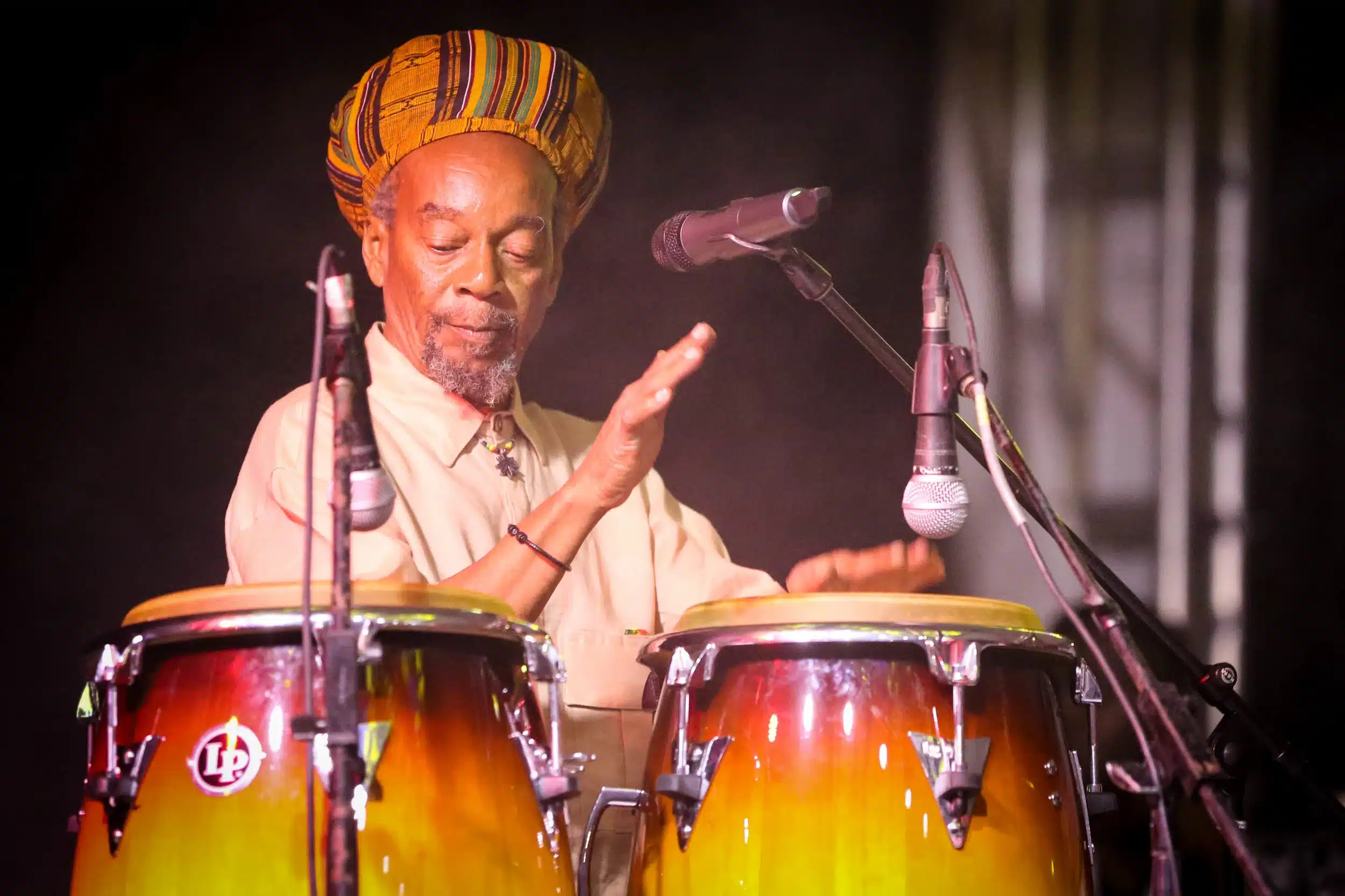 An older man wearing a colourful hat plays two conga drums on stage, surrounded by microphones, with dramatic lighting highlighting his focused expression.