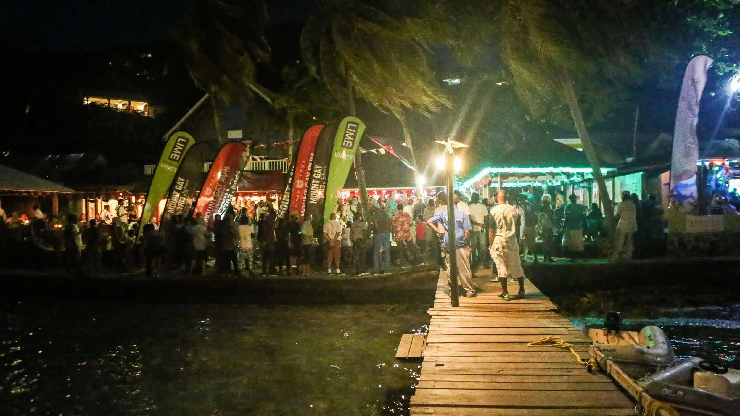 A lively nighttime waterfront party with a crowd gathered near colourful flags and fairy lights. Palm trees sway in the background, and people are walking on a wooden jetty leading to the festivities.