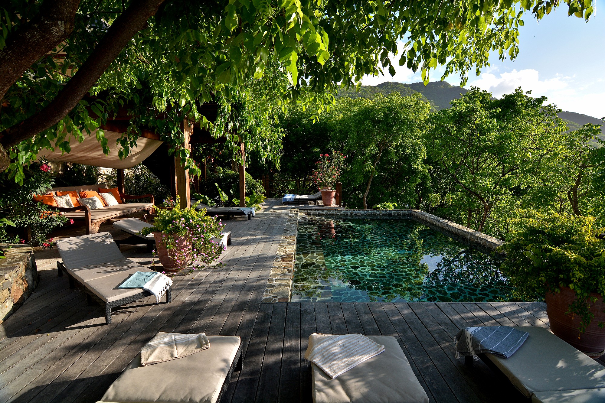 A serene outdoor lounge at Cassava Lower Bay Bequia features cushioned chairs and sofas surrounding a clear stone-edged pool, shaded by lush green trees and overlooking distant hills under a bright sky. Photo